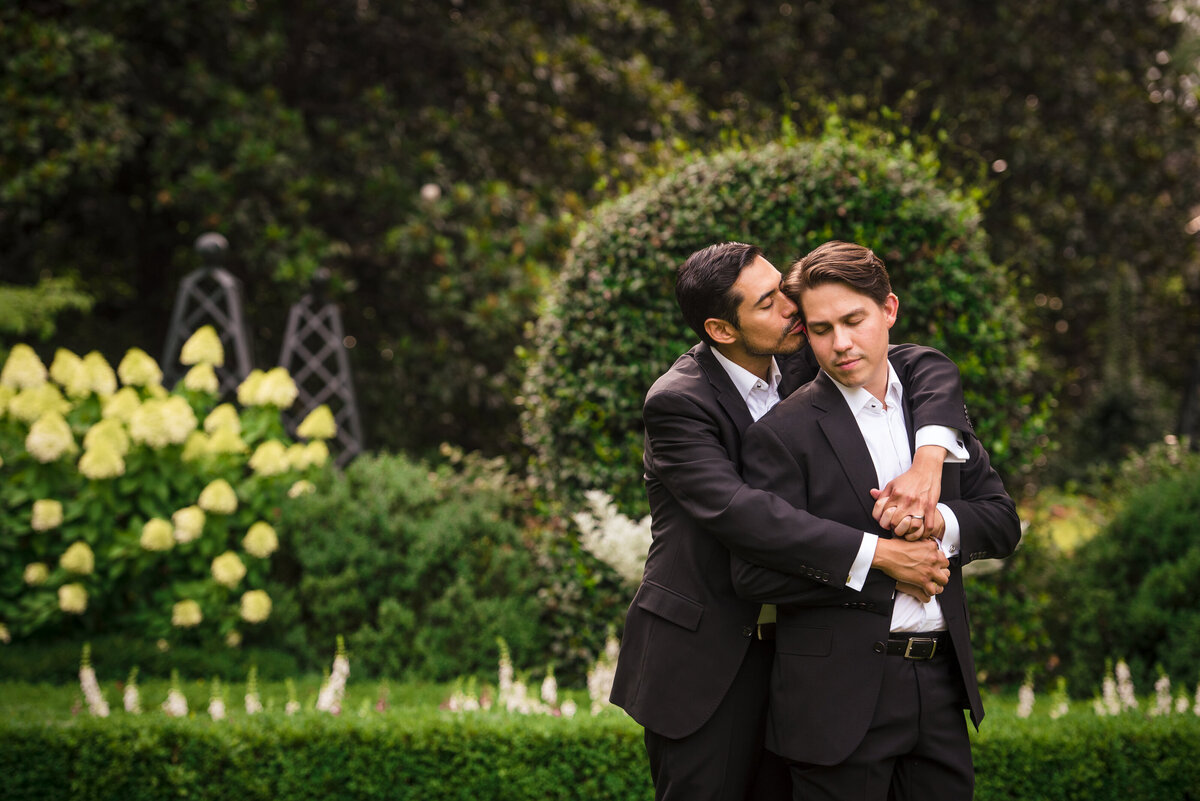 Portrait-of-a-gay-couple-their-arms-are-wrapped-around-each-other-in-a-spoon-position-and-one-groom-is-kissing-the-other-groom-on-the-temple-in-the-garden-of-The-Duke-Mansion