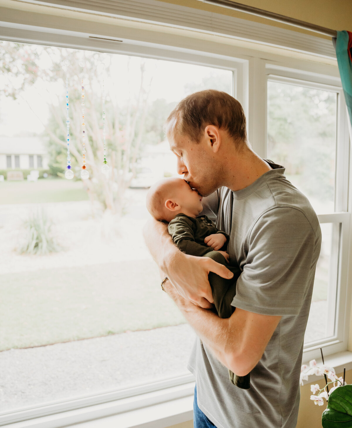 Newborn Photographer, a father stands near a window with his baby, and gives him a kiss