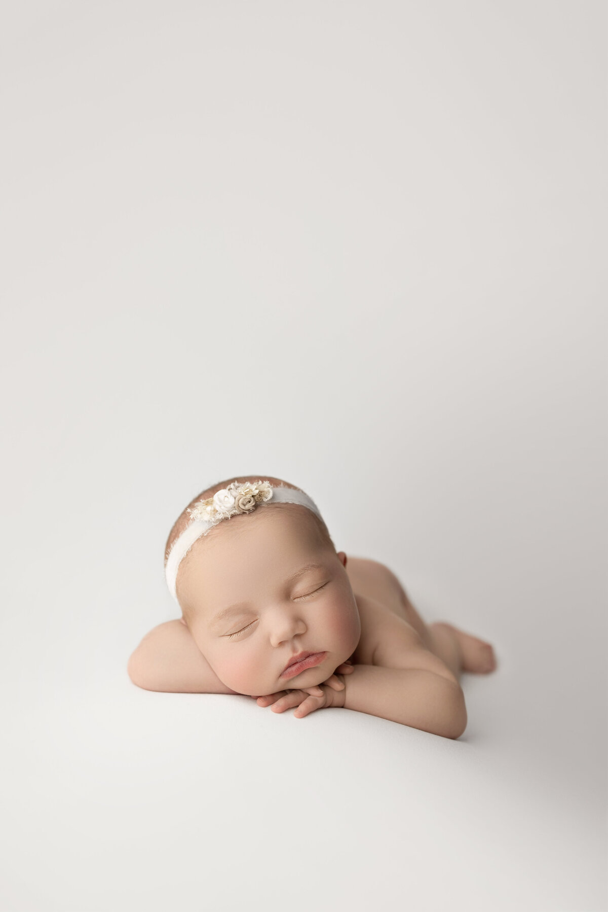 Fine art newborn baby photoshoot captured by best Main Line newborn baby photographer Katie Marshall. Newborn baby girl is sleeping on her belly on white stretch fabric. Baby girl's hands are folded underneath her chin and her head resting on her forearm. She is wearing a delicate floral headband.