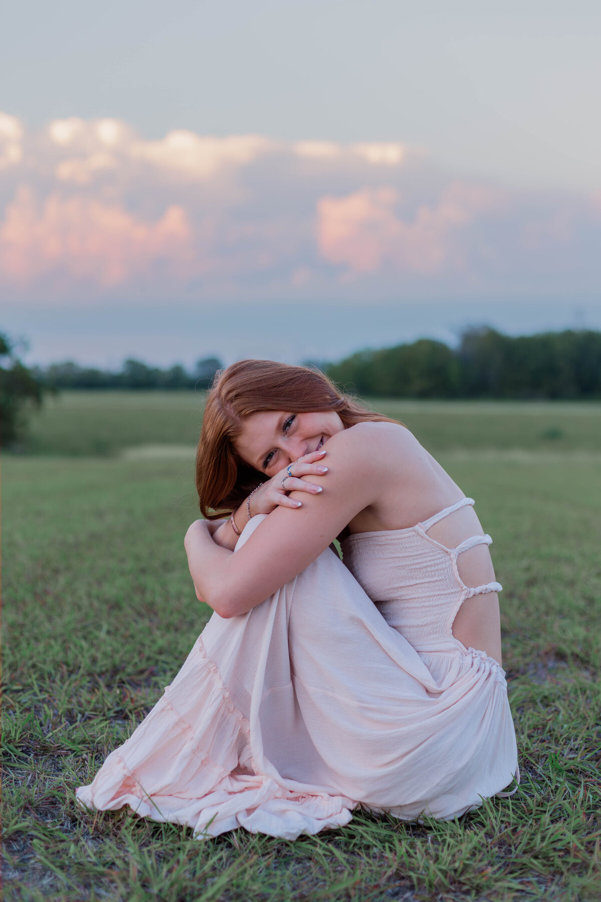 A shy girl smiles as she sits on the ground hugging her knees in a field at sunset