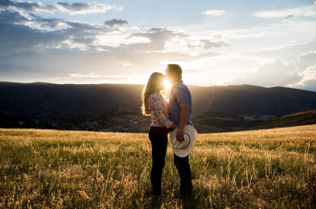 A man kisses his fiancée's forehead while he holds a cowboy hat  standing in a golden field at sunset, taken by Denver engagement photographer Casey Van Horn.