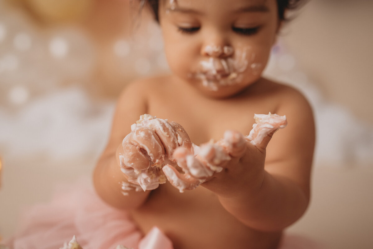 one year old baby girl at atlanta ga cake smash portrait studio wearing a pink tutu and smashing and eating a cake  with pastel rainbow balloon garland behind her