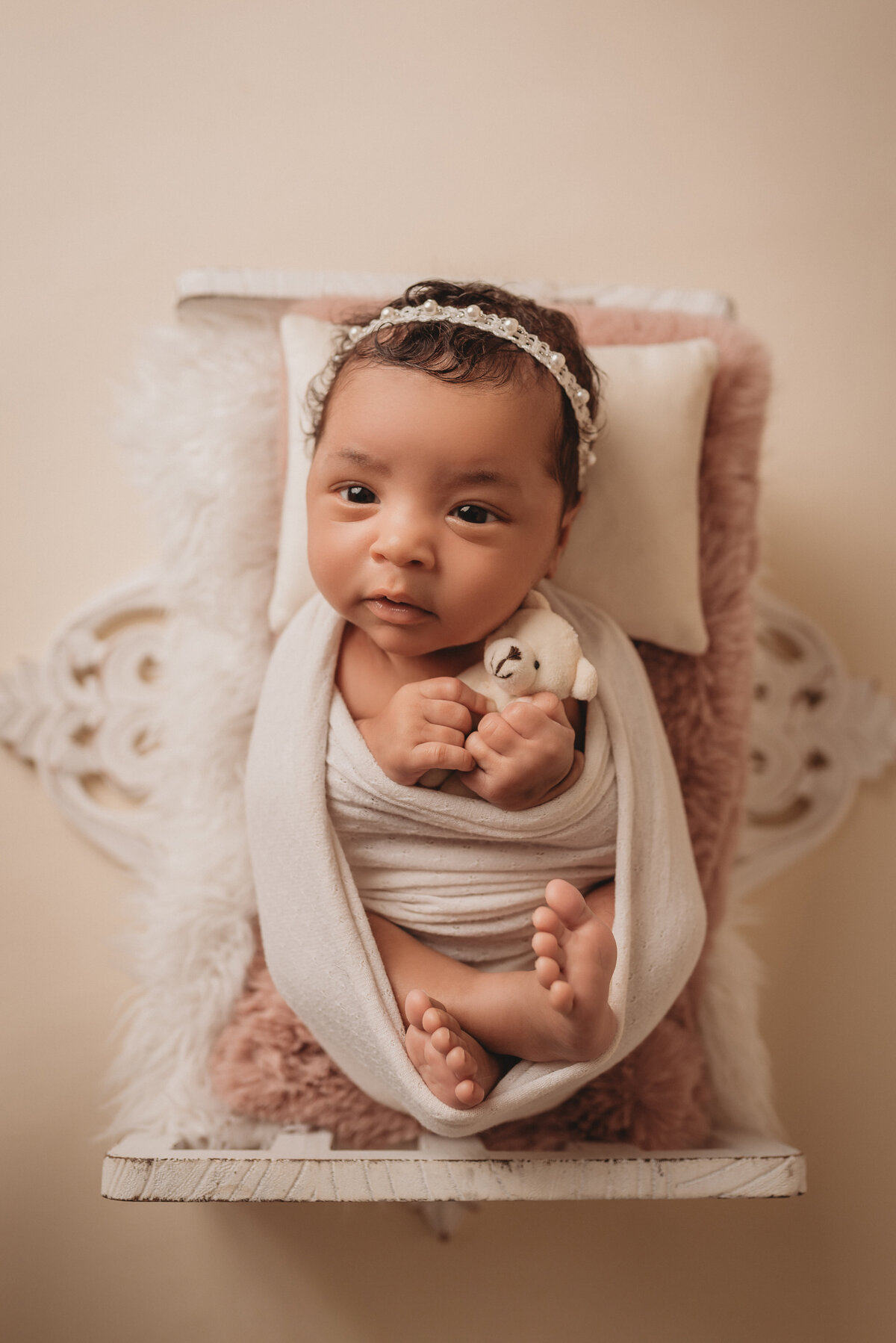 Newborn photography portrait session with baby girl wrapped in cream fabric with fingers and toes showing, laying in tiny white bed holding a teddy bear and looking at the camera