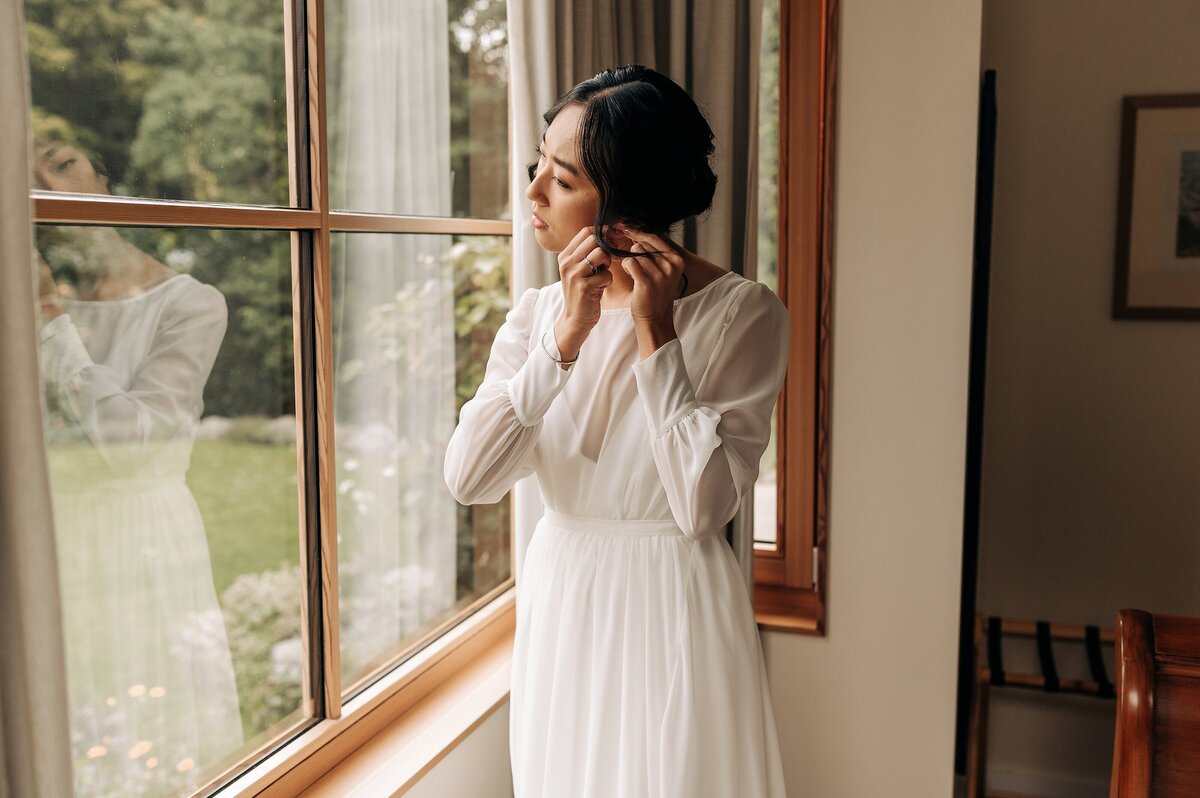 bride putting on earrings in wedding dress by wooden bay window in character home airbnb christchurch getting ready for wedding accommodation