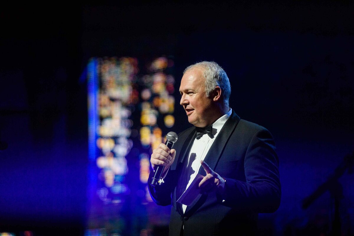 Man holds mic up to face dressed in tuxedo on stage for dinner entertainment at destination event