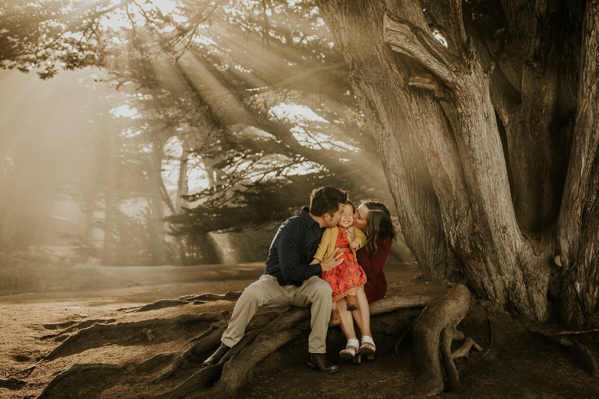 Portrait of parents kissing toddlers cheeks while sitting in a wooded area with sunbeams coming through