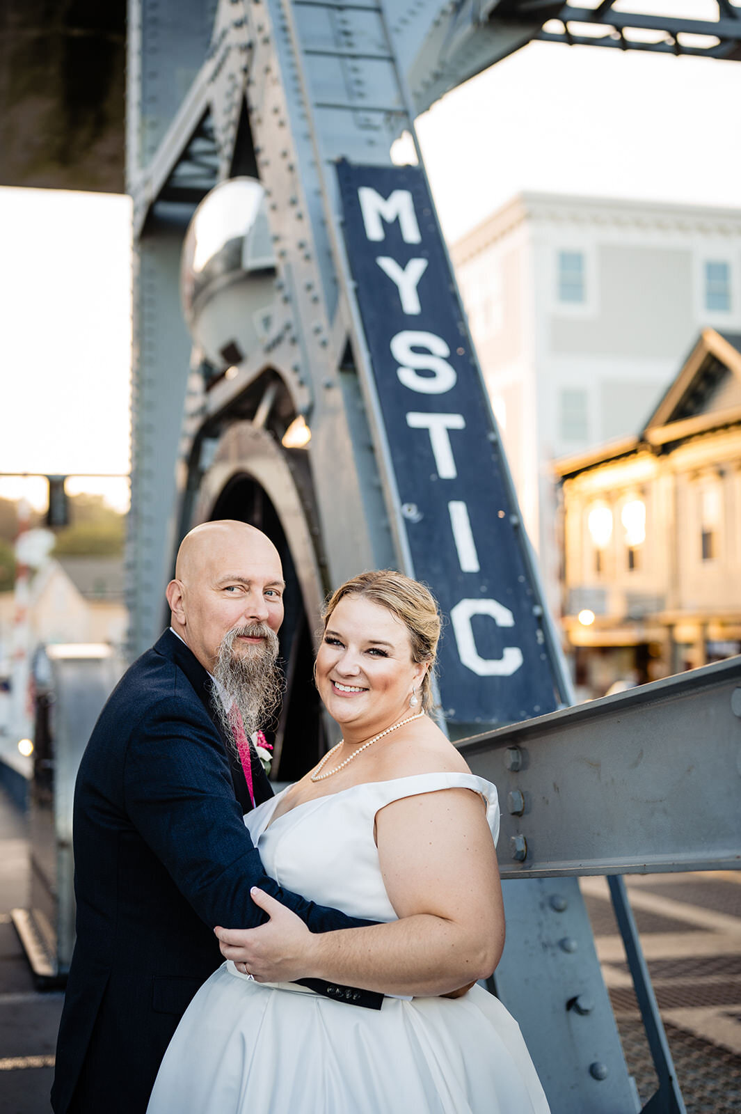 A smiling bride and groom stand close together in front of the iconic Mystic drawbridge, with the name "Mystic" clearly visible