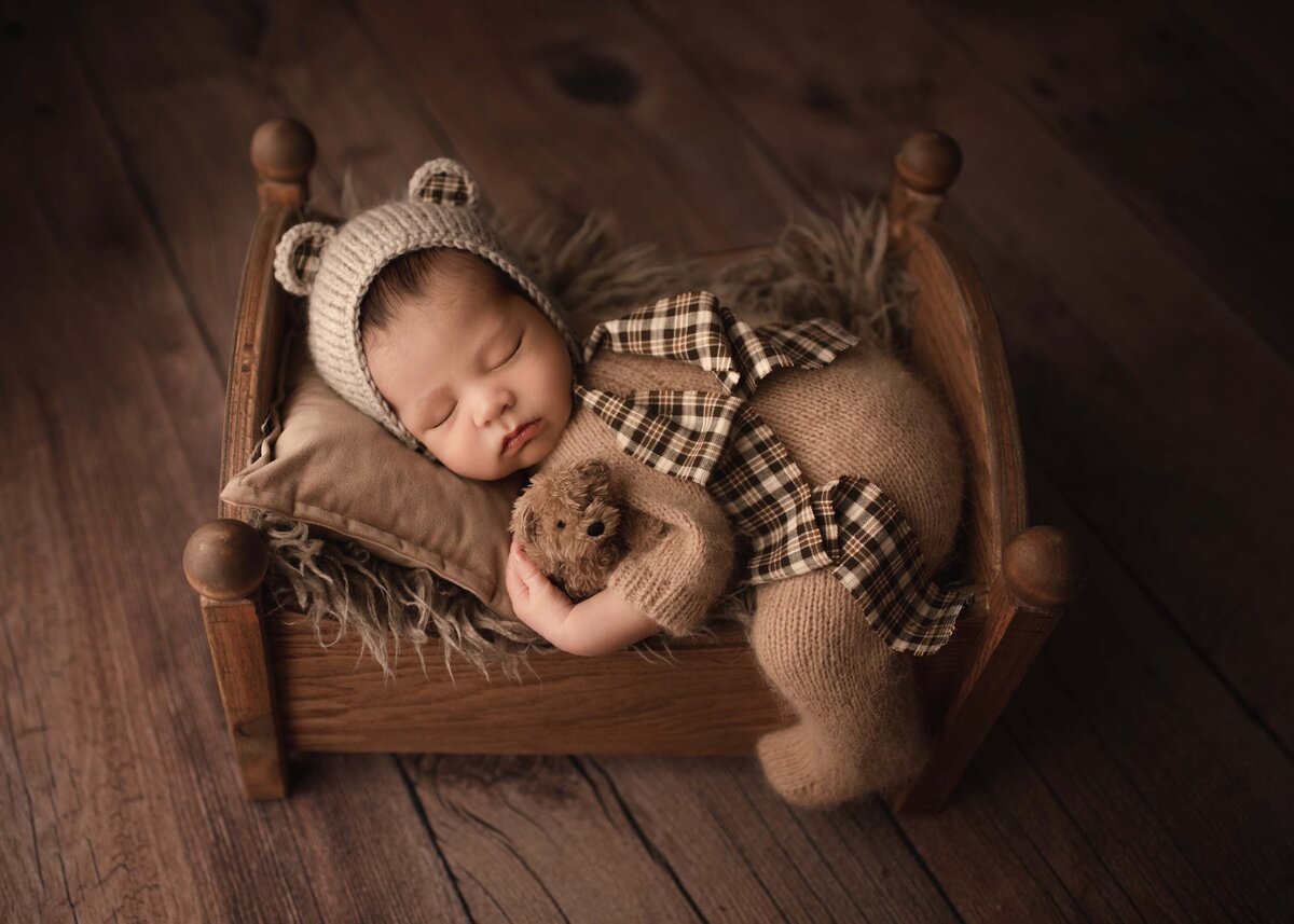 Baby boy wearing bear bonnet, holding tiny bear posed on a bed prop
