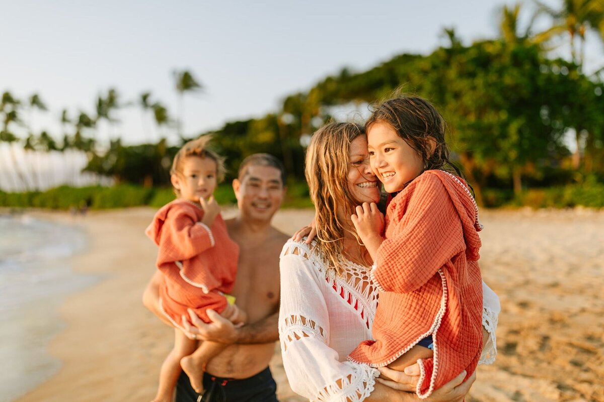 A mother and father, each hold their child wearing swimsuits walking along the beach in Maui.