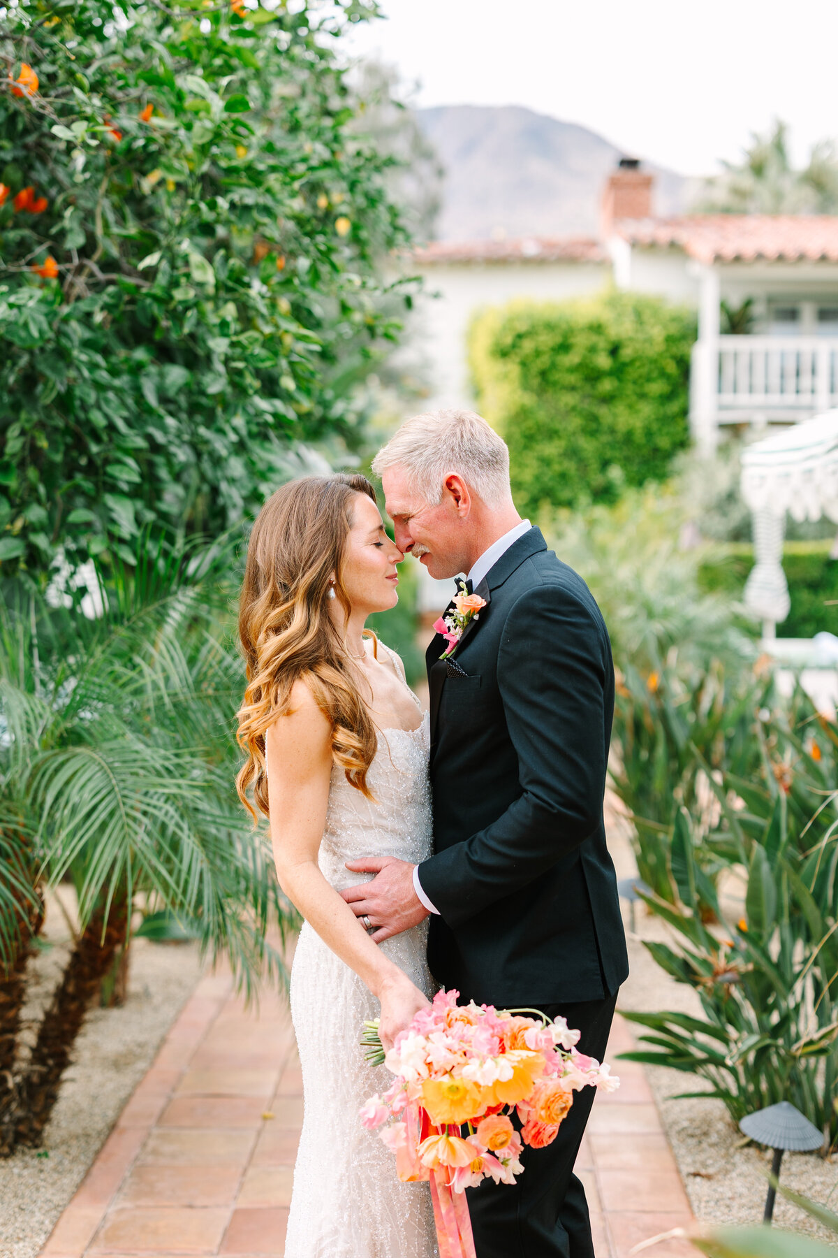 bride and groom embracing outside under the trees while bride holds orange and soft pink bridal bouquet.