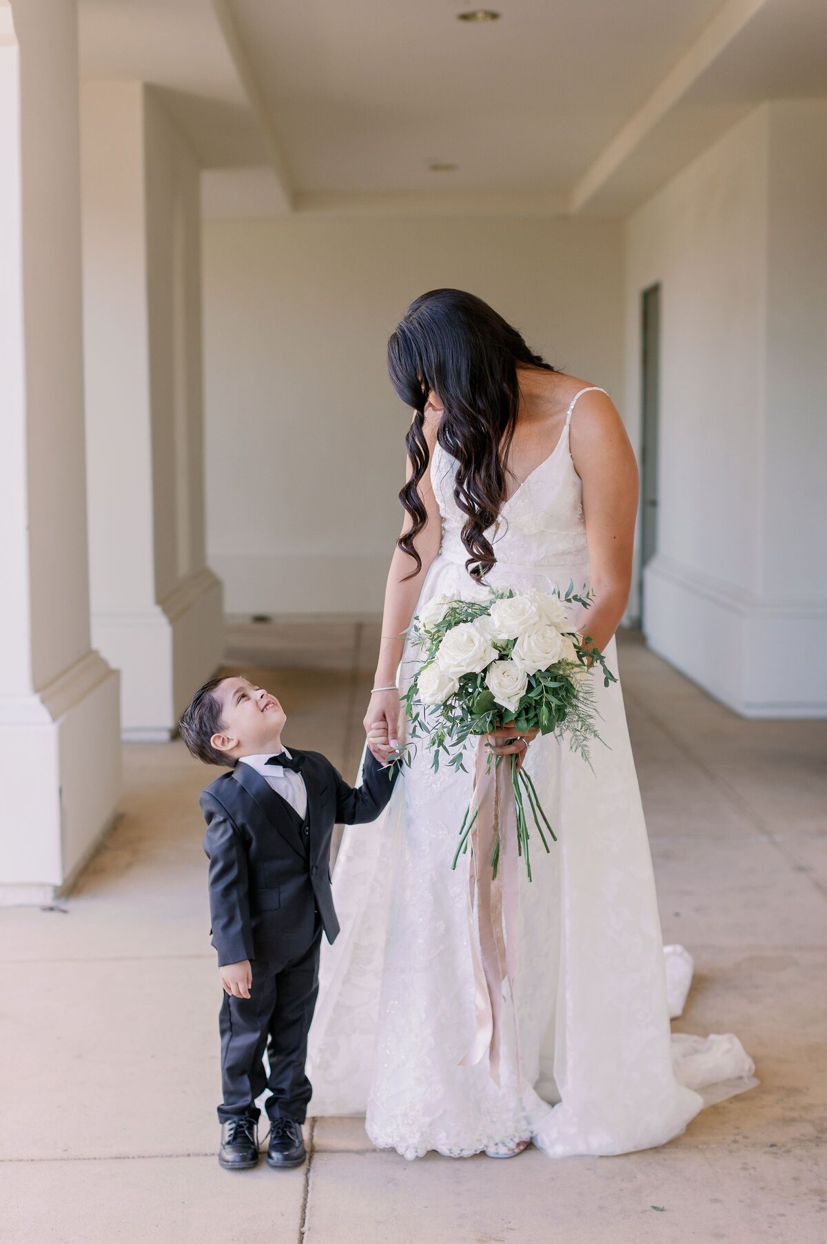 This little man is wearing a lovely suit standing next to the bride who holds a large white rose bouquet.