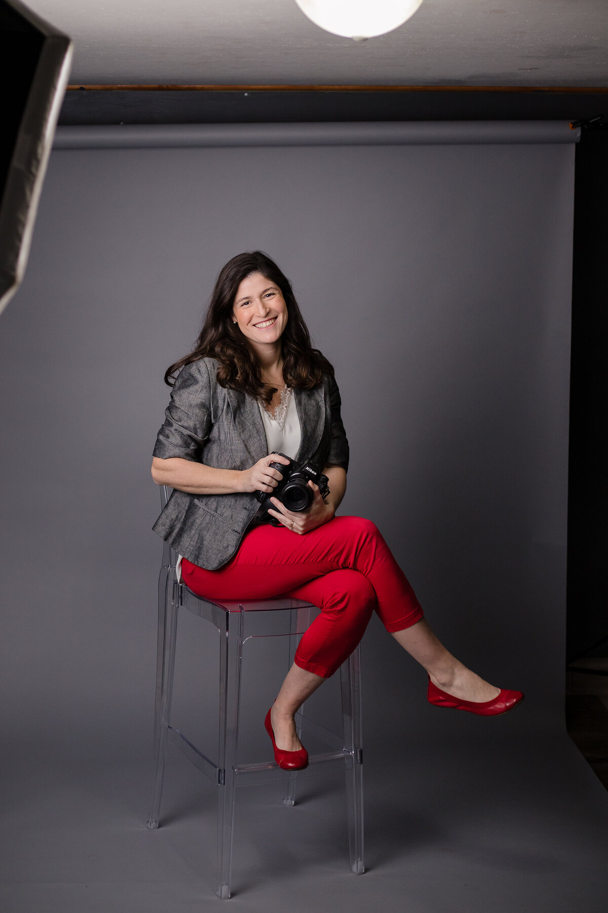 photographer Lori pickens in red pants and red shoes sitting on clear chair in photo studio with flash shown in corner