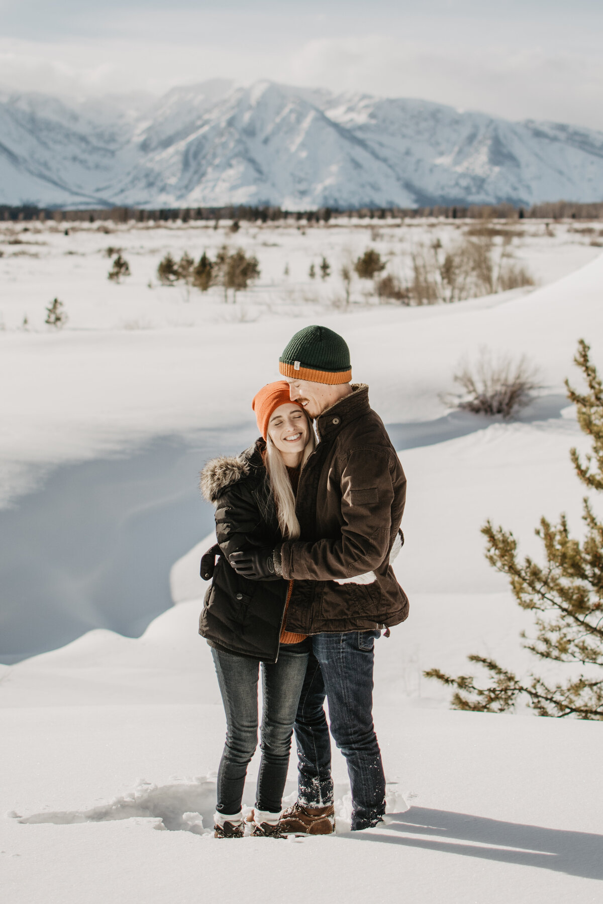 jackson hole photographers captures standing in the snow, a young engaged couple holds each other as the man kisses the woman's forehead in Jackson Hole for their winter engagement session
