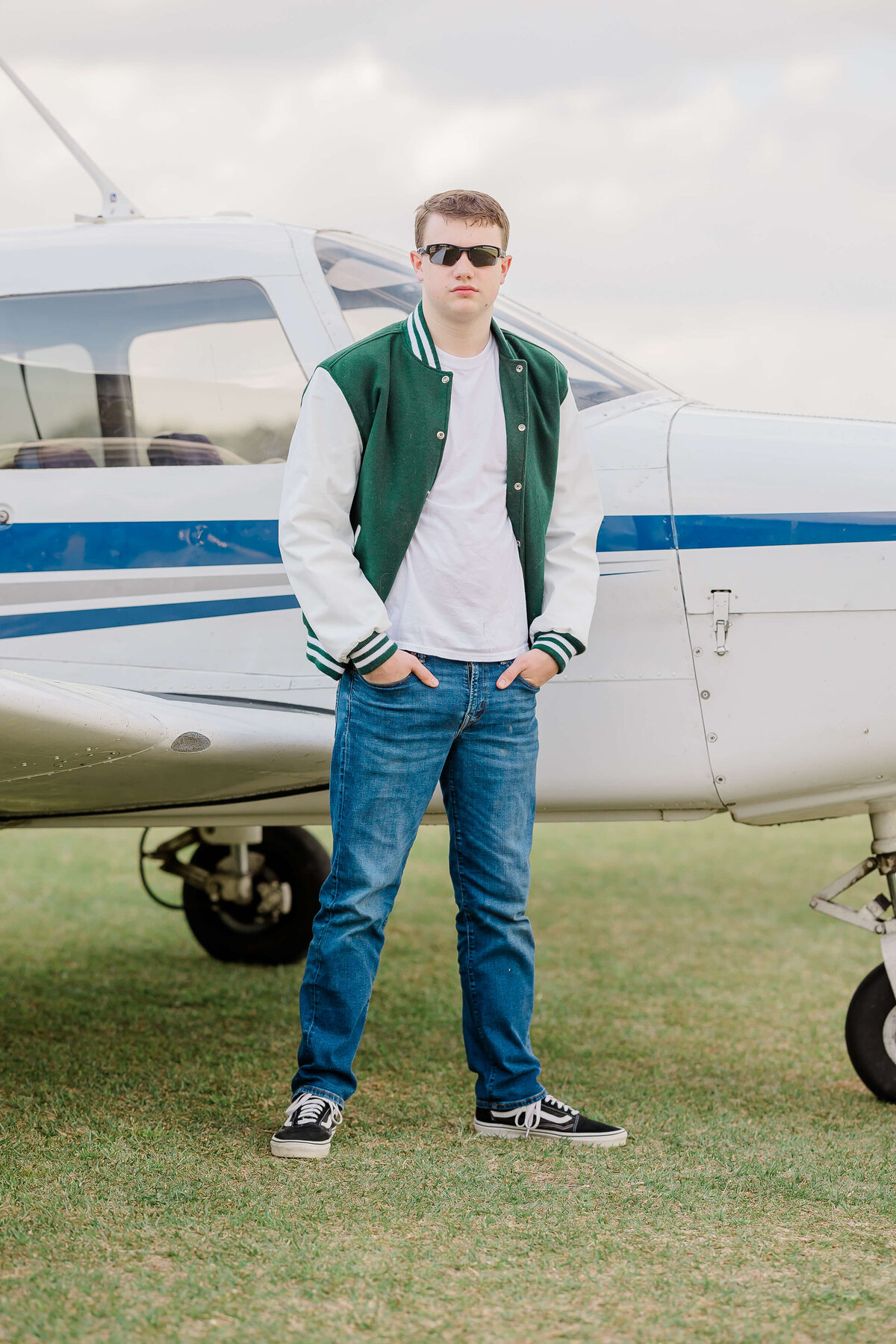 A person wearing sunglasses, a green and white varsity jacket, jeans, and black sneakers is standing in front of a small white and blue airplane on a grassy area. Their hands are in their pockets.