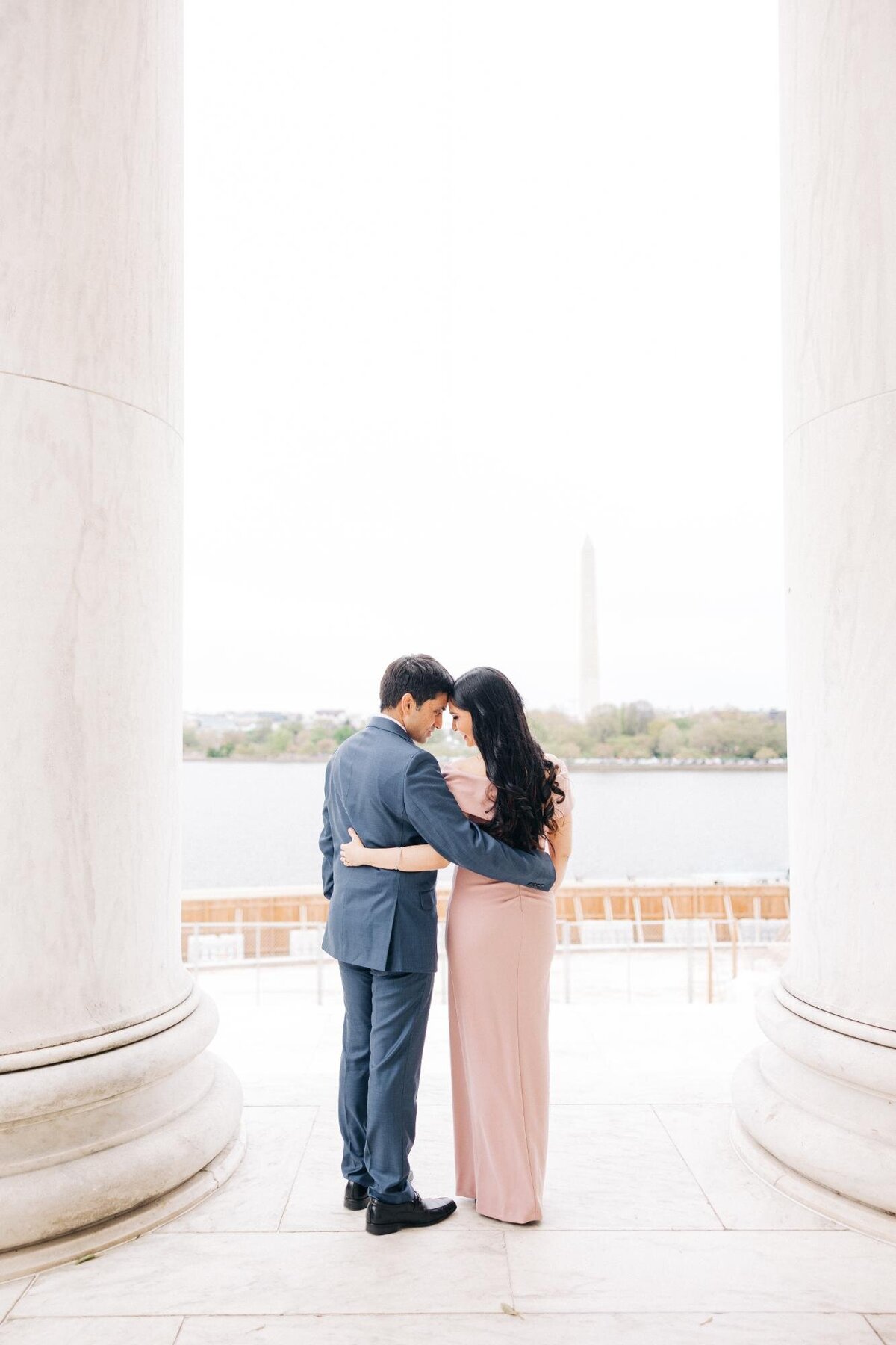 A couple embracing under a white columned structure with the washington monument visible in the background.