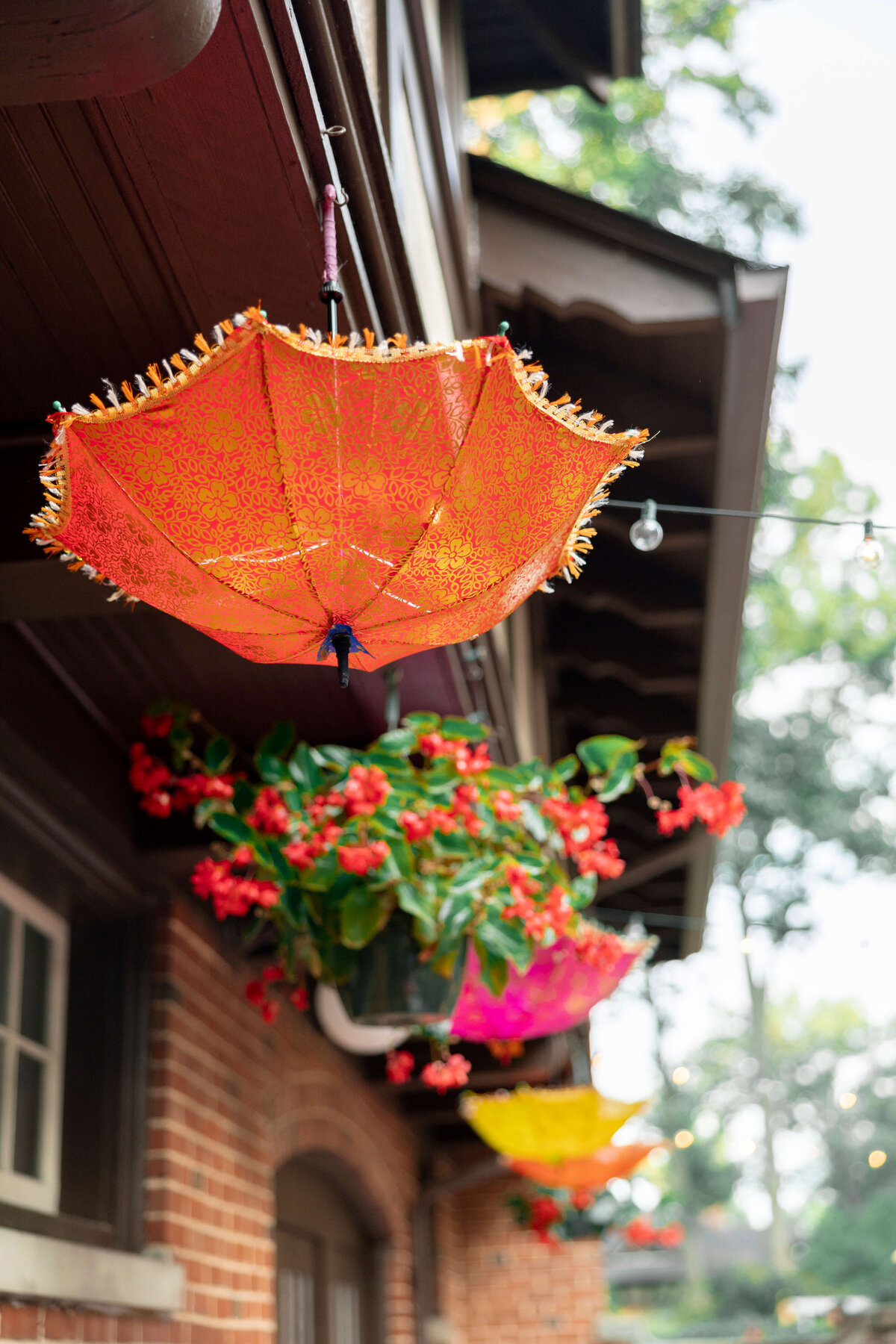 Upside-down orange umbrella with decorative fringe hangs from a building adorned with red flowers in window boxes. More colorful umbrellas hang in the background, creating a festive display.