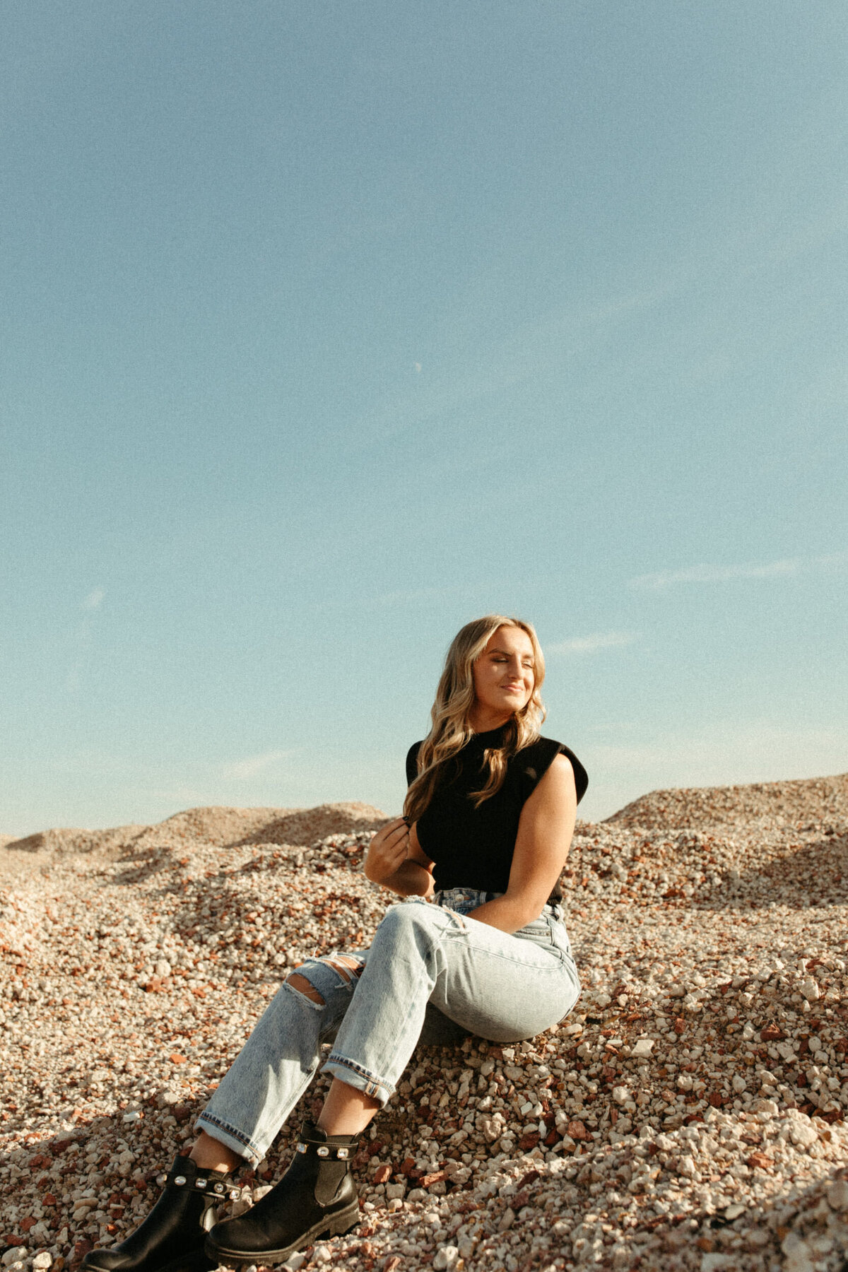 High school senior sitting on top of gravel mound with blue sky behind her