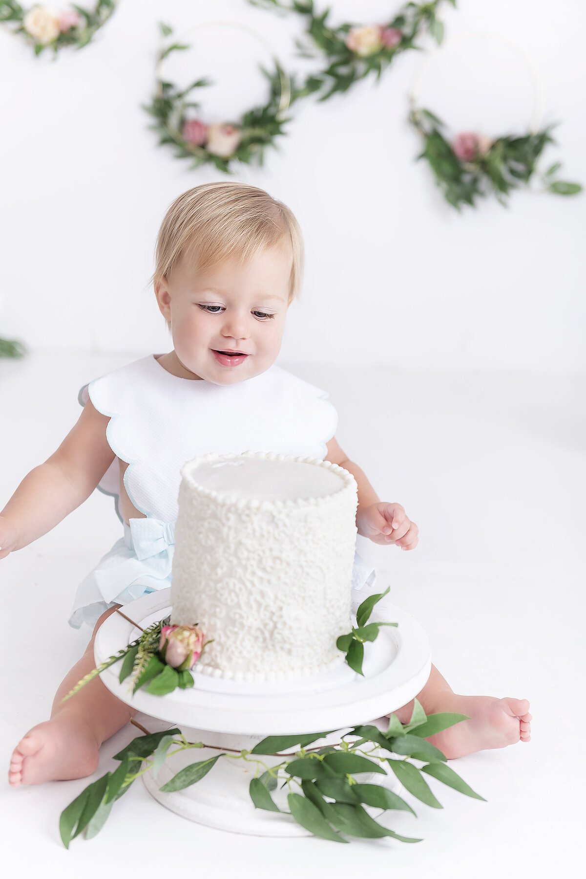 A toddler in a white dress looks at a cake she was about to smash in a studio