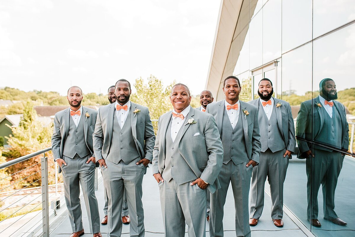African American groom in a light gray suit with a white shirt and orange bow tie stands with his hands in his pockets in front of his groomsmen who are dressed in light grey suits with white shirts and orange bowties on the balcony of The Liff Center in Nashville next to a mirrored wall at the Nashville Opera for a Nashville Wedding at Noah Liff.