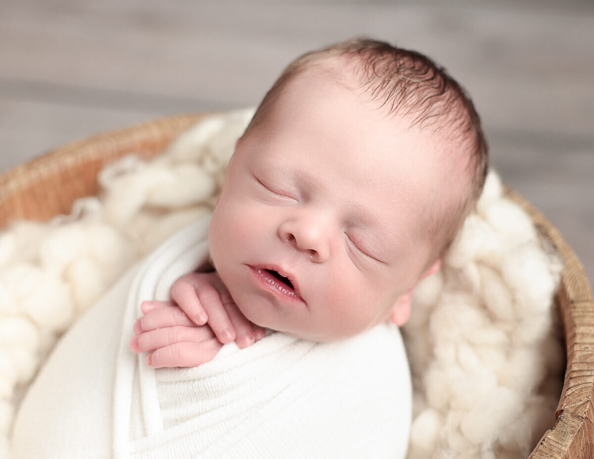 A peaceful newborn boy, swaddled in a soft white blanket, nestled in a cozy basket at a photography studio in Rochester, NY. The image captures the innocence and tranquility of newborn photography, highlighting the delicate features of the baby.