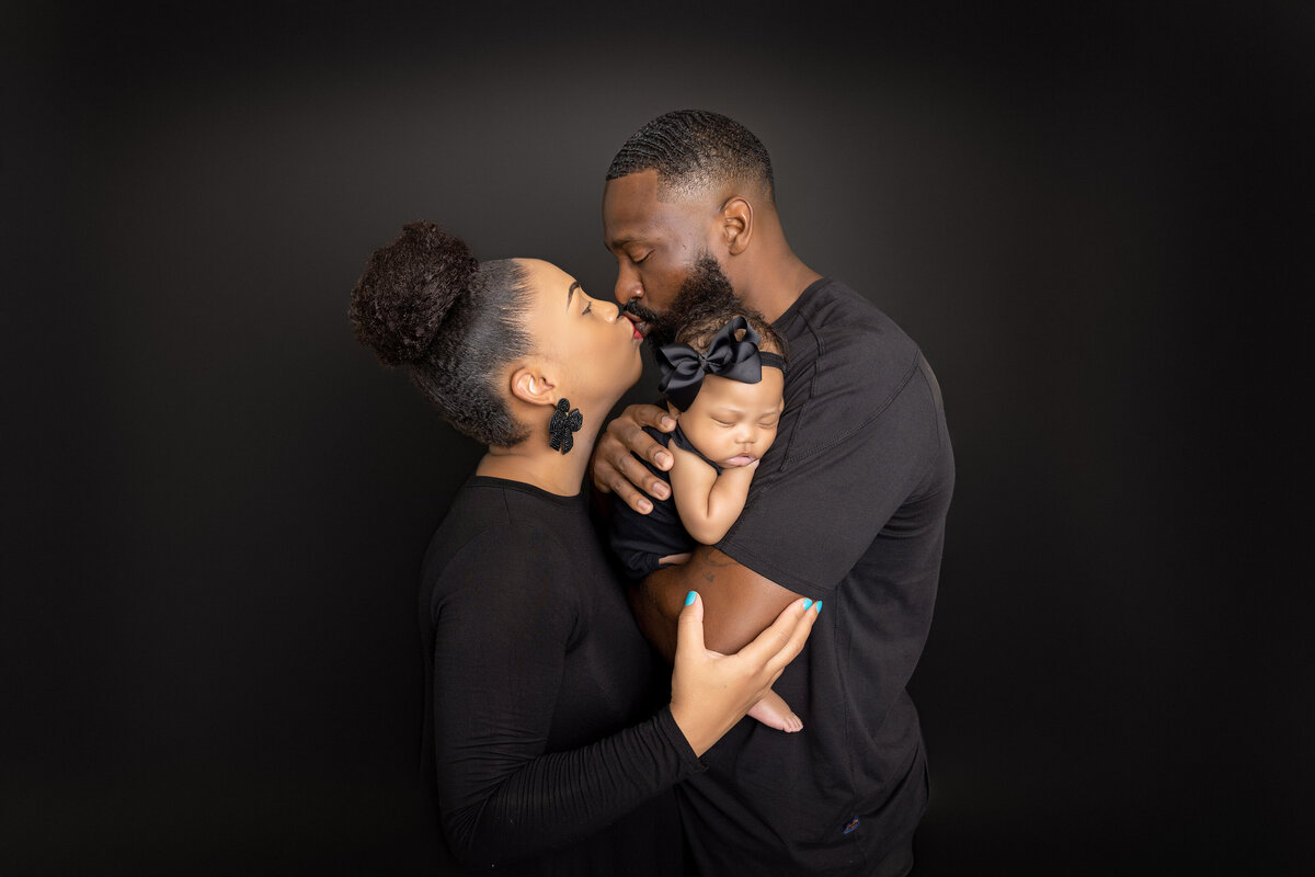 A Jacksonville newborn photographer captures a tender family portrait: a man cradles his sleeping baby in a black outfit with a matching bow, while a woman lovingly kisses his cheek. The dark background beautifully highlights their expressions.