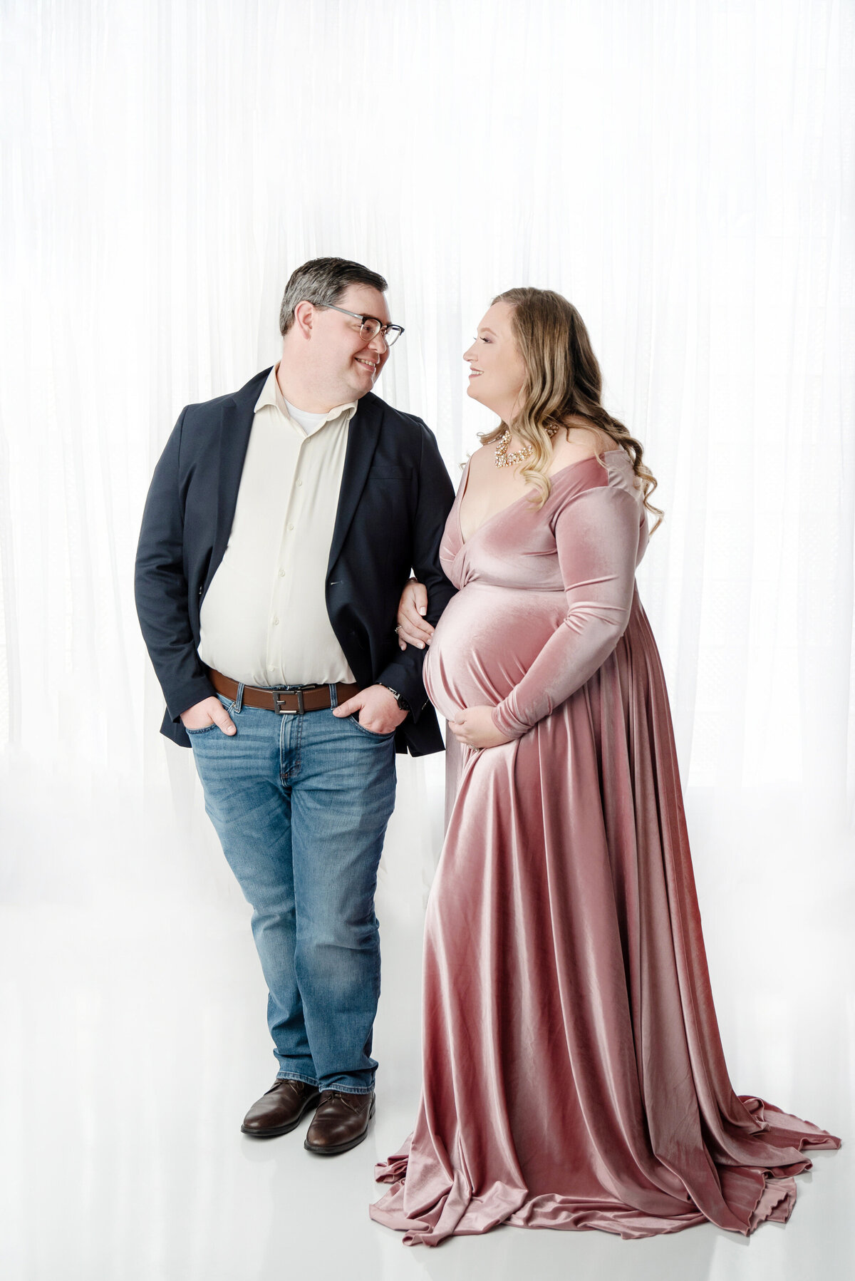 st-louis-maternity-photographer-expecting-mother-and-father-linking-arms-against-white-background-he-in-a-casual-suit-she-wearing-a-pink-velvet-gown