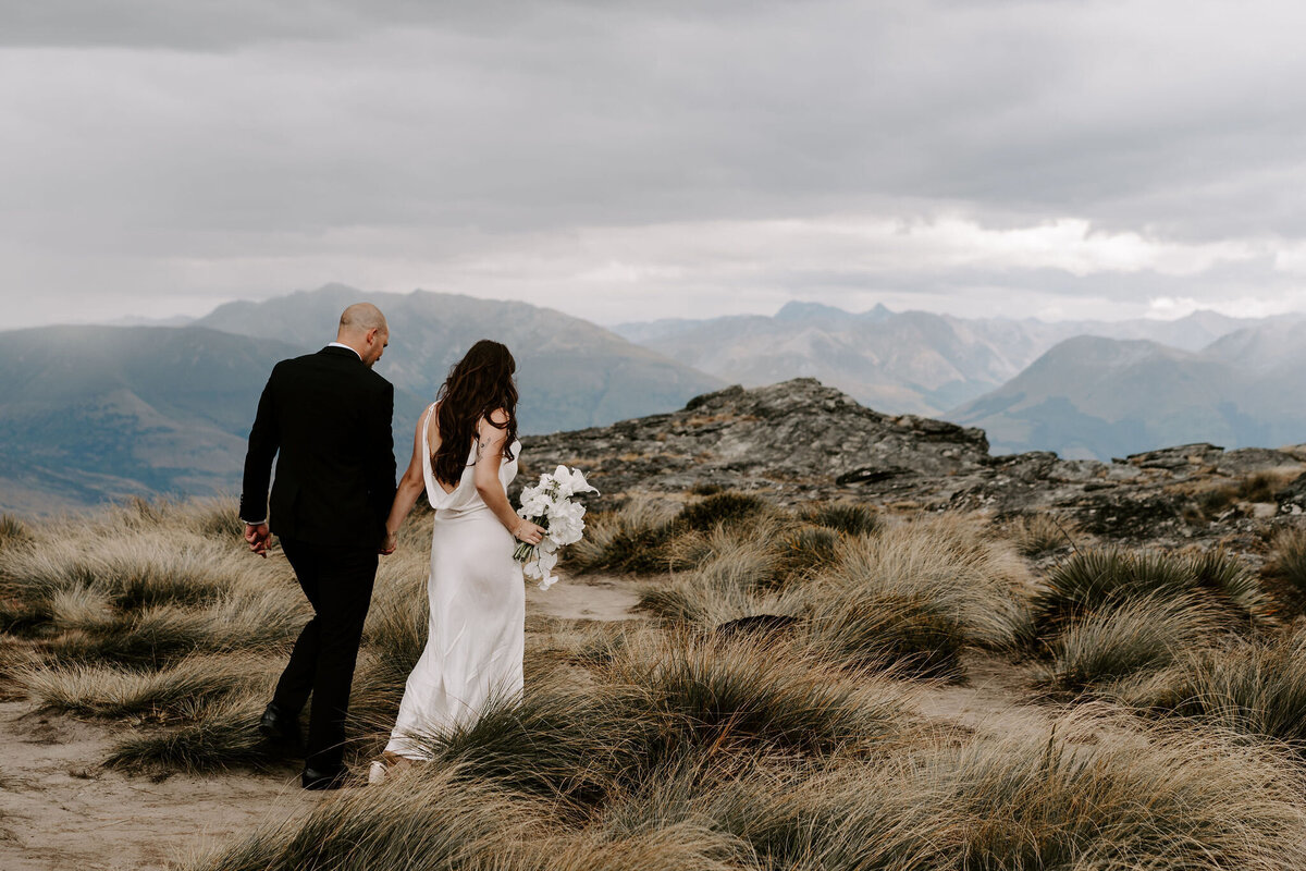 The Lovers Elopement Co - bride and groom walk through long grass on mountain with lake below, heli wedding