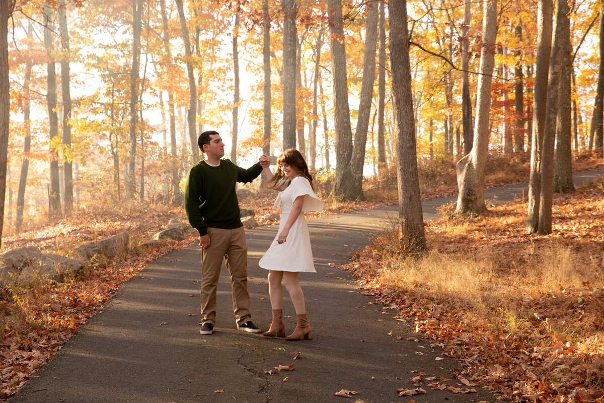 A man in a dark blue sweater twirls his fiance in a white dress on a path with autumn leaves orange trees Harriman State park fall engagement session New York wedding photographer