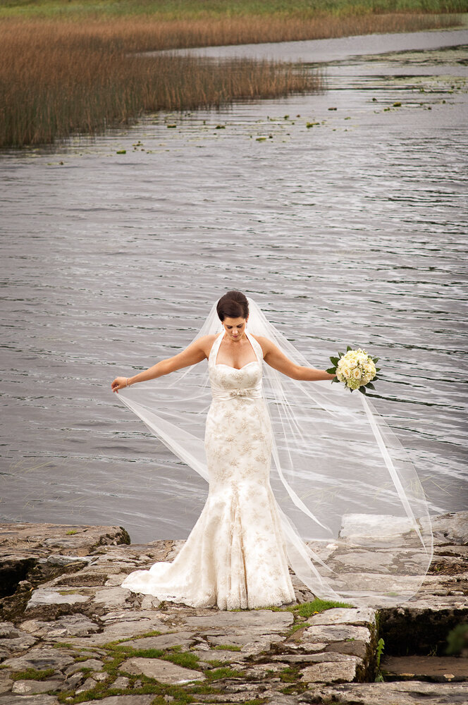 brunette bride wearing a lace, trumpet style, halter neck wedding dress and long veil standing at lake in Killarney