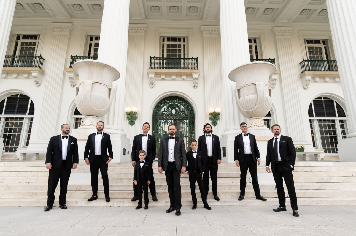 A groom in a black tuxedo stands with his groomsmen in front of a grand building with large white columns, reflecting timeless style and sophistication.
