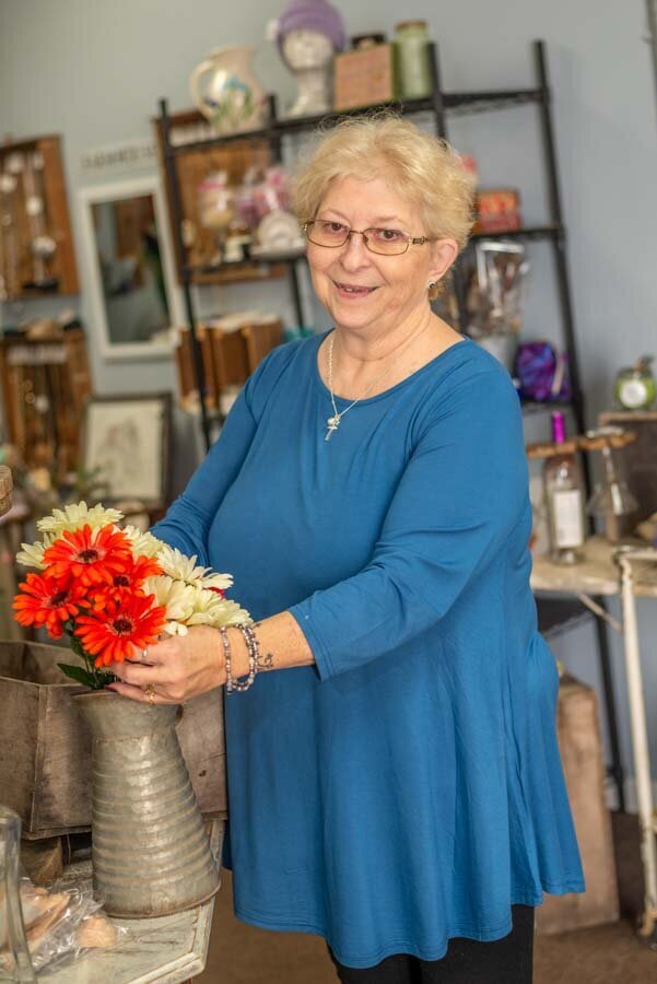 Elderly woman in a blue top smiling while arranging colorful flowers in a metal vase inside an antique shop.