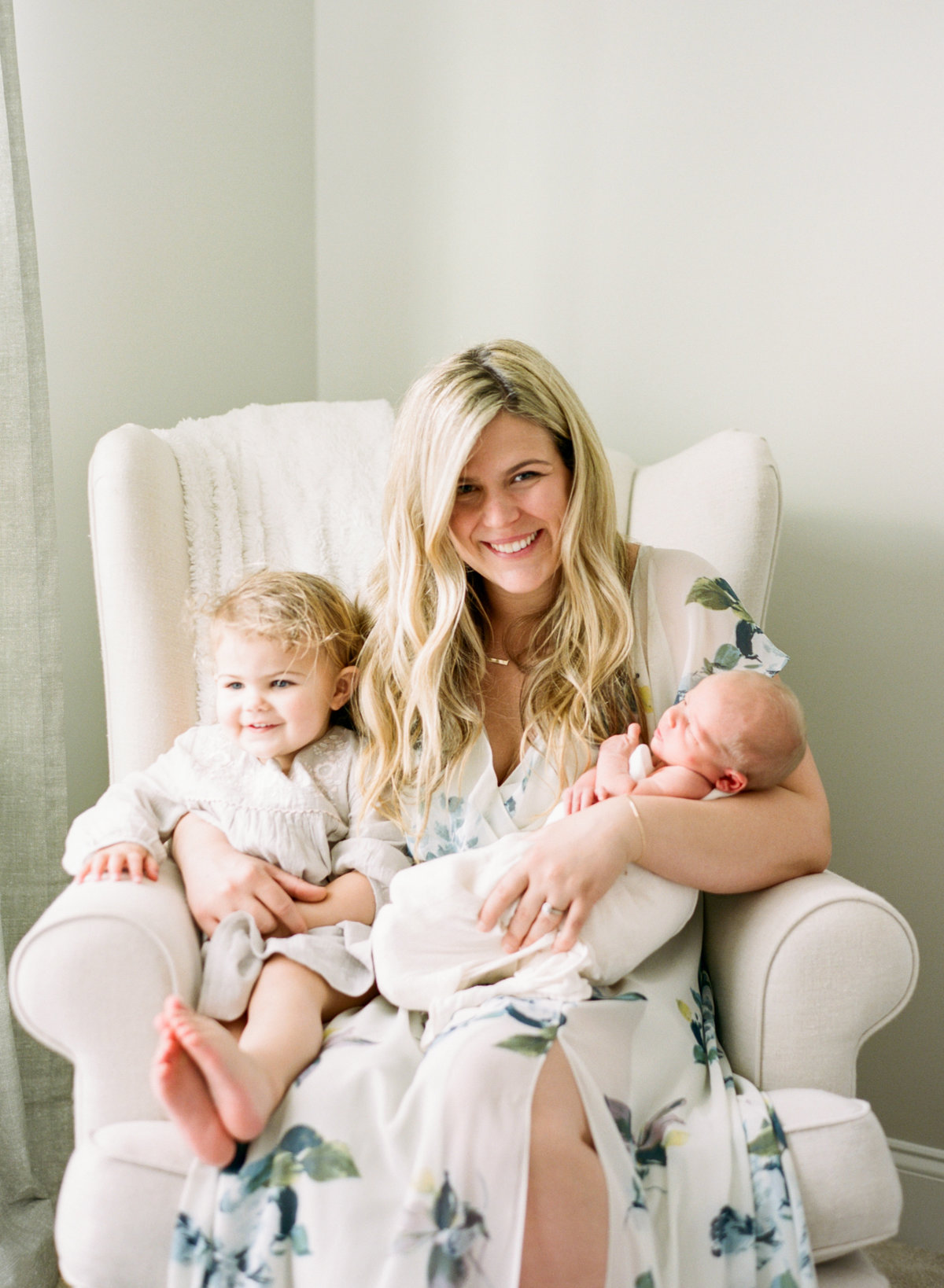 Mom sits in a rocker and smiles while holding her newborn and toddler during a Raleigh newborn session. Photographed by Raleigh Newborn Photographer A.J. Dunlap Photography.