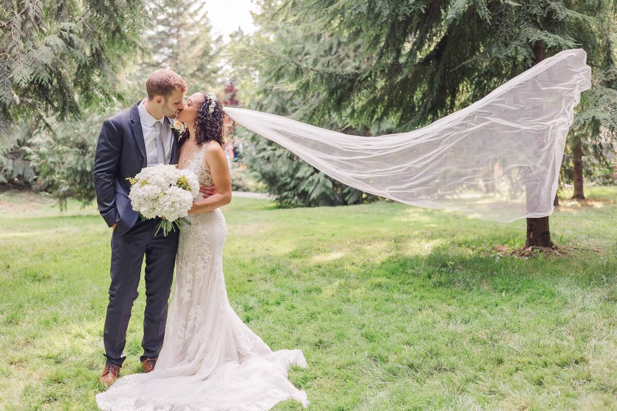 Bride and groom, in an embrace, kissing in the middle of a forest, while the brides long veil flies in the wind