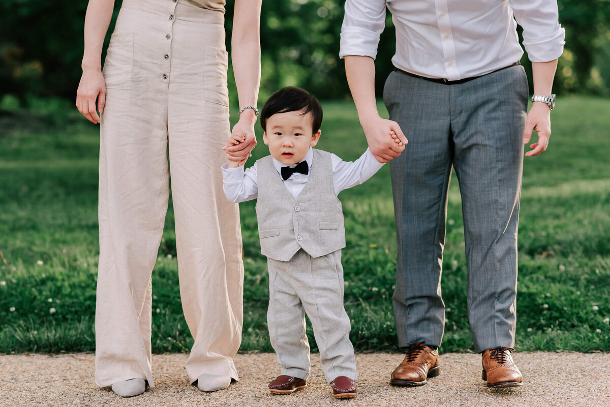 A baby in grey slacks, vest and black bowtie, standing between his parents