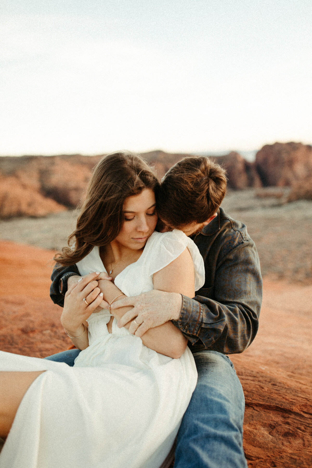 Guy snuggling his fiancé as they sit on a rock in the desert with cliffs in the background