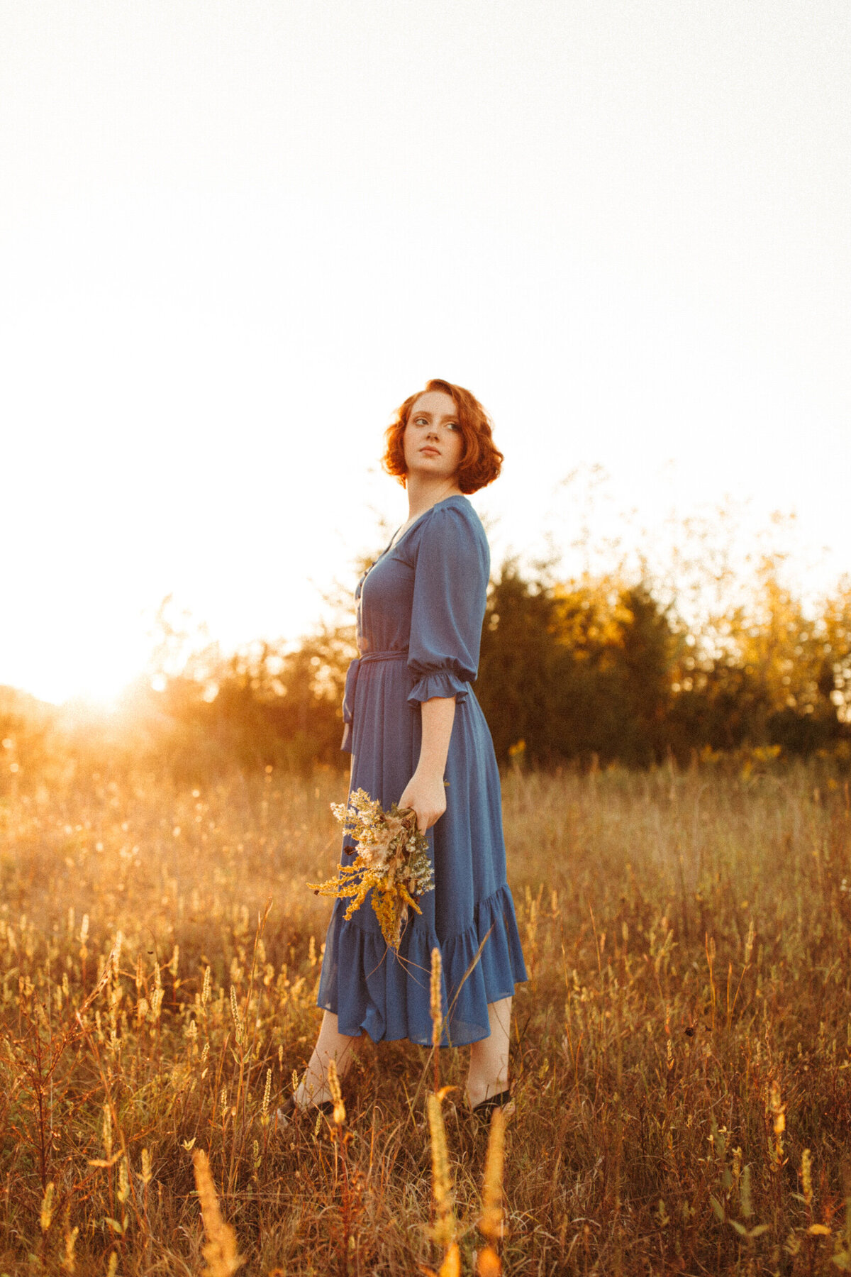 High school senior in blue dress holding a bundle of wildflowers and walking in a field