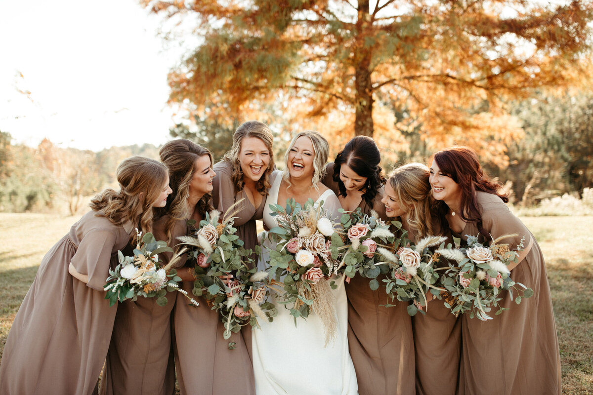 Bride laughing with her six bridesmaids holding floral bouquets on a fall day