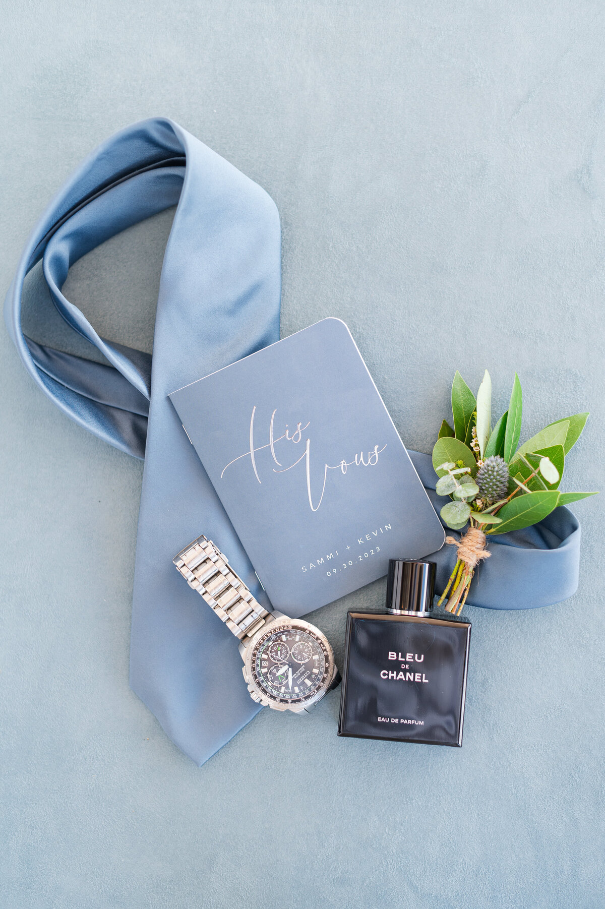 Flat lay of groom's tie, blue vow book, watch, cologne bottle and boutonniere on a blue suede background.
