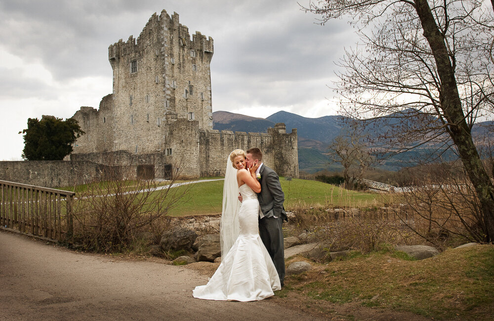 blonde bride wearing a mermaid style wedding dress and long veil being kissed by her husband wearing a dark grey tailcoat wedding suit while standing in front of Ross Castle, Killarney