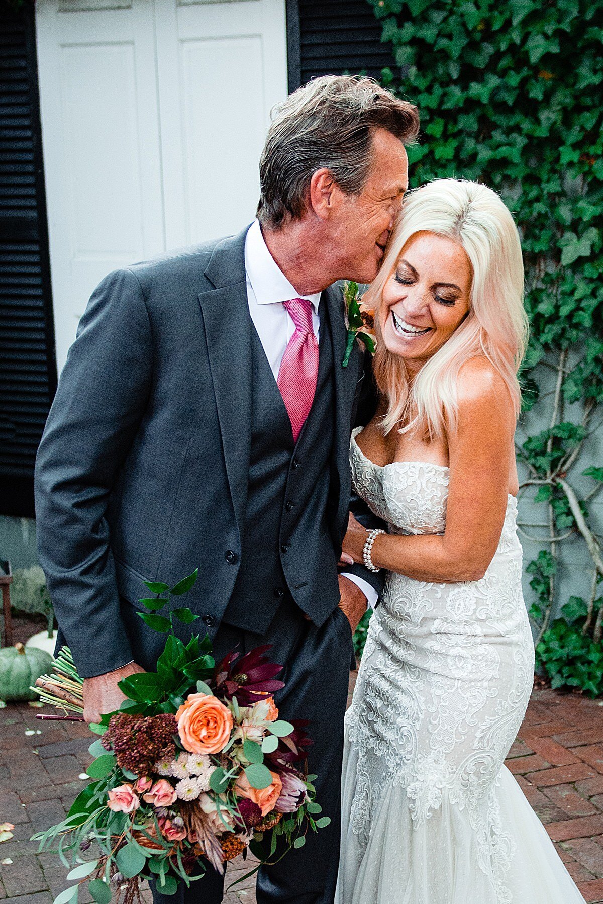 The groom, wearing a three piece navy suit and pink tie with a white shirt holds the brides bouquet as he whispers in her ear. The bride, looking down at the bouquet, laughs.  The bride is wearing a strapless mermaid style wedding gown with lace detailing on the bodice. The skirt is sheer organza. They are standing in an ivy covered courtyard with a white door. The bridal bouquet is full of burgundy, peach, pink, blush and ivory flowers.