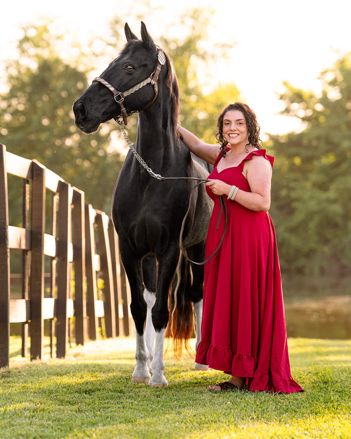 black haired female equestrian in a flowy red dress standing next to her black tobiano spotted saddle horse in a field. behind them is a lake and beautiful golden hour light behind them