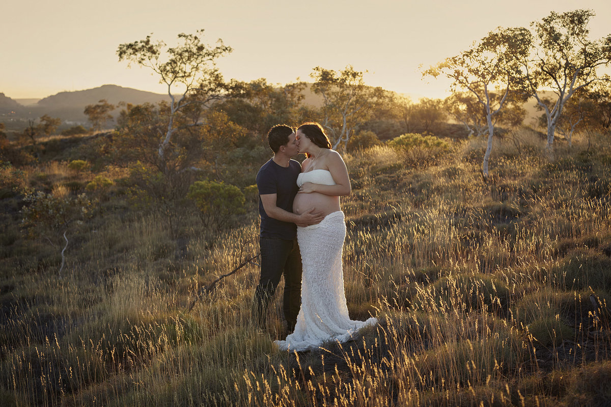 Pregnant wife and husband kissing and cuddling each other in natural bushland with golden sunlight