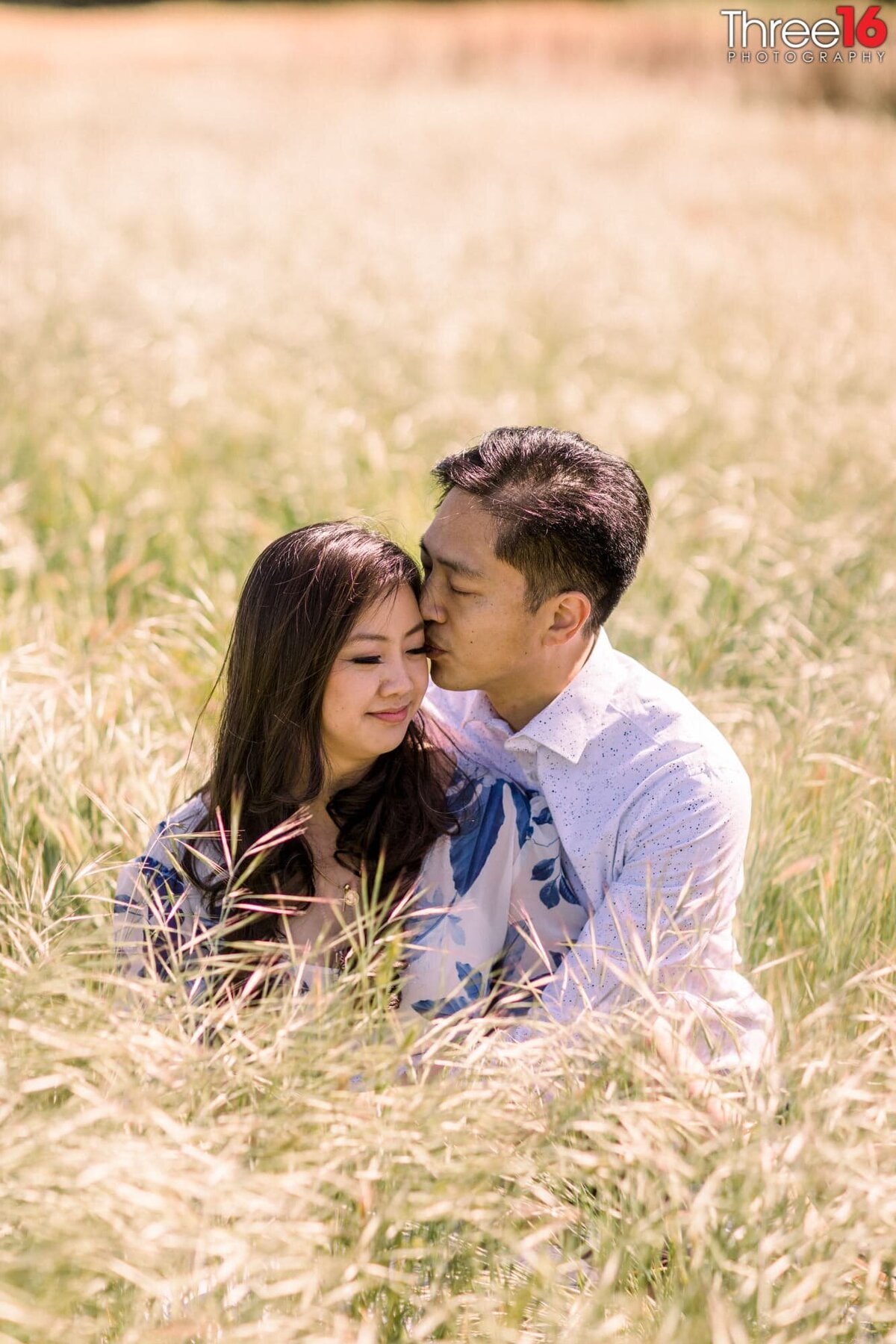 Groom to be kisses his Bride's cheek as they sit in tall grass for the engagement session