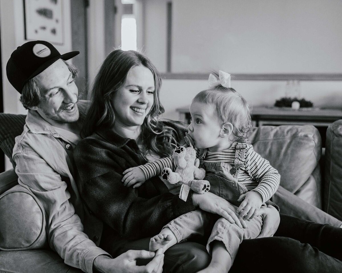 Maddie Rae Photography black and white family picture. dad is leaning on the arm of the couch, mom is leaning on him and baby is on moms lap. mom and dad are smiling at baby and baby is looking at them