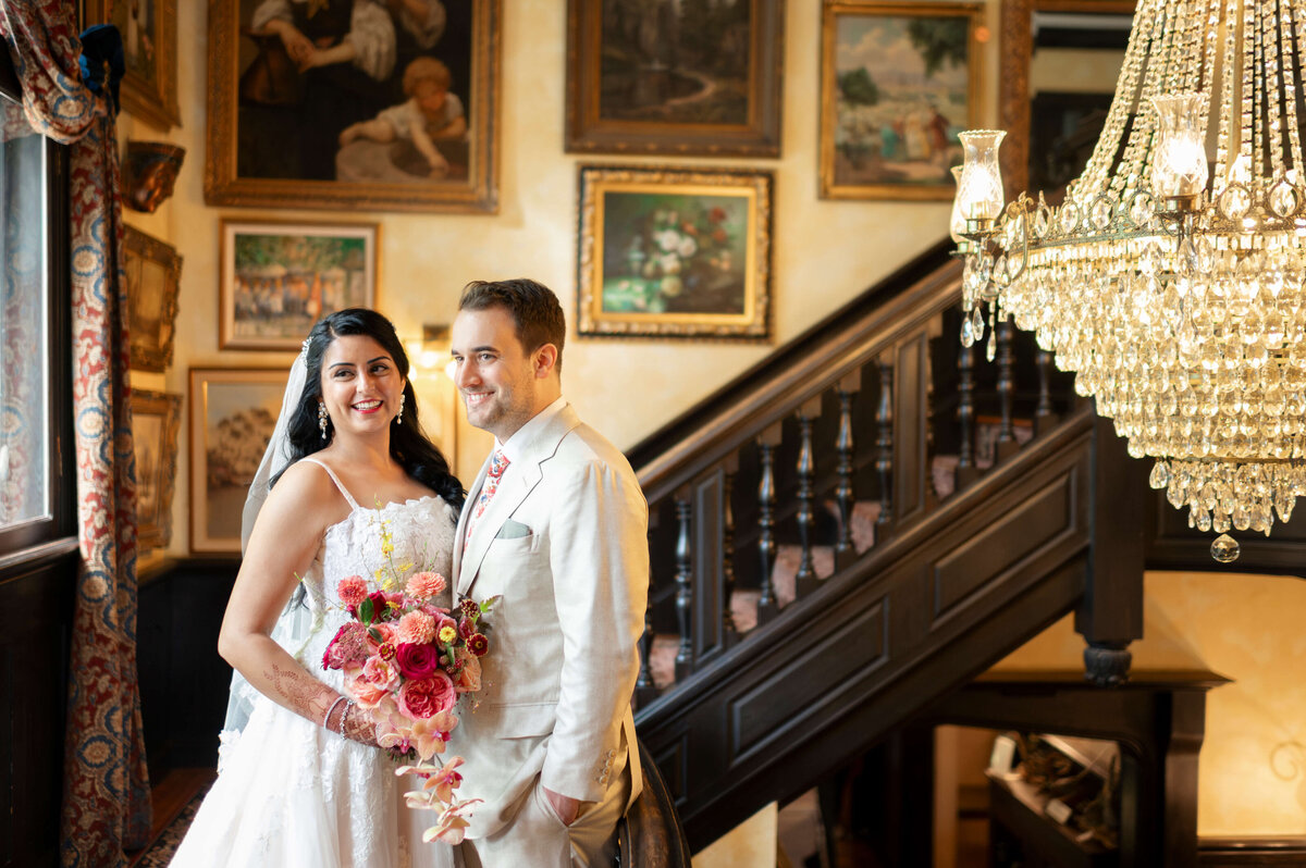 A bride and groom smile, posing in an elegantly decorated room with a grand staircase, ornate chandelier, and artwork on the walls. The bride holds a bouquet of pink and red flowers.