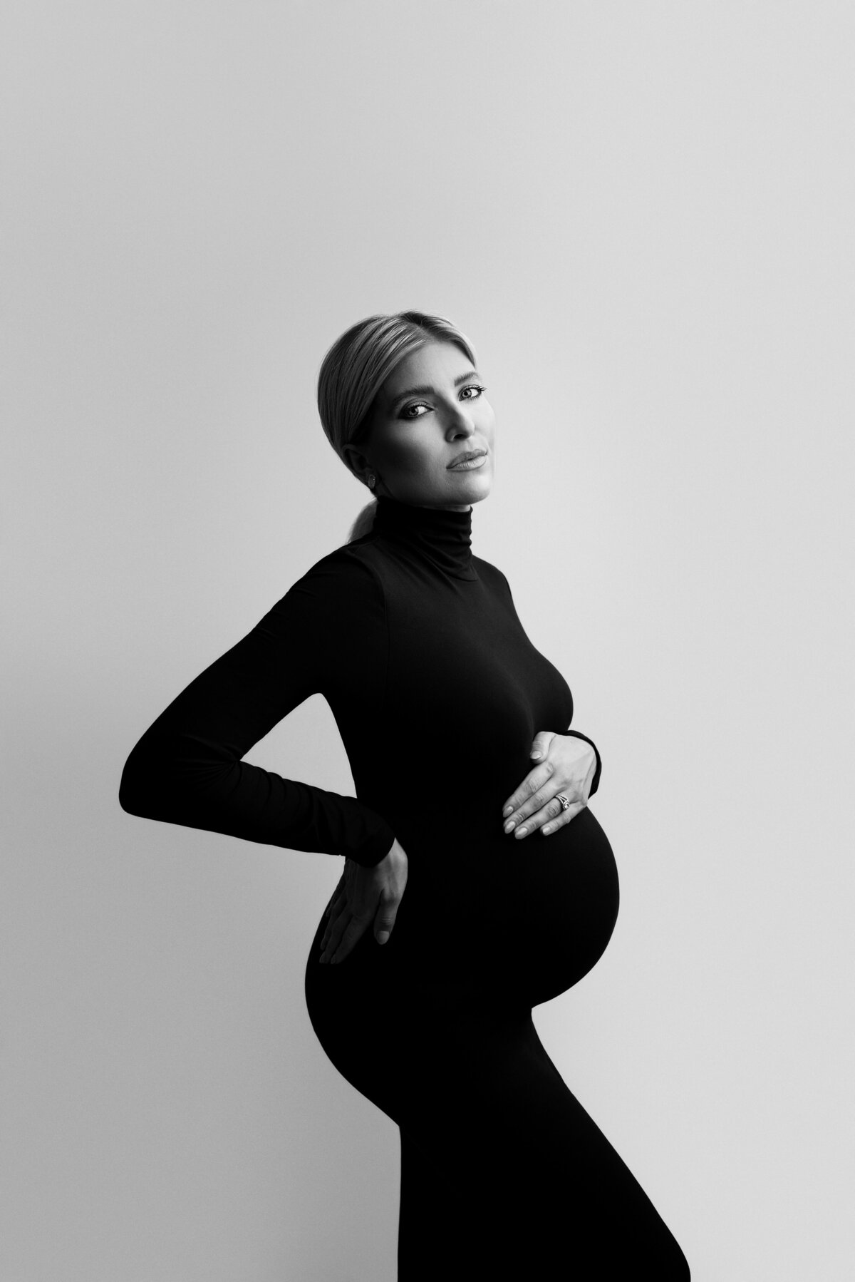 Woman poses for a fine art maternity photoshoot with Philadelphia Main Line's best maternity photographer Katie Marshall. Woman is standing side profile to the camera in a long, black body con dress.  Her forearm is resting on the small of her back, her back arm is resting atop of her baby bump. She is looking over her shoulder with a closed-mouth smile. Her hair is tied back in a low ponytail.