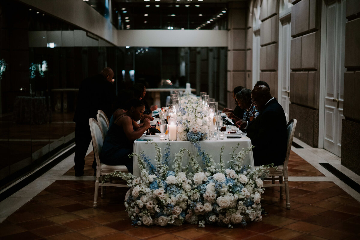 Elegant dining scene with people seated at a candlelit table adorned with lush white and blue flowers, creating a warm, intimate atmosphere.