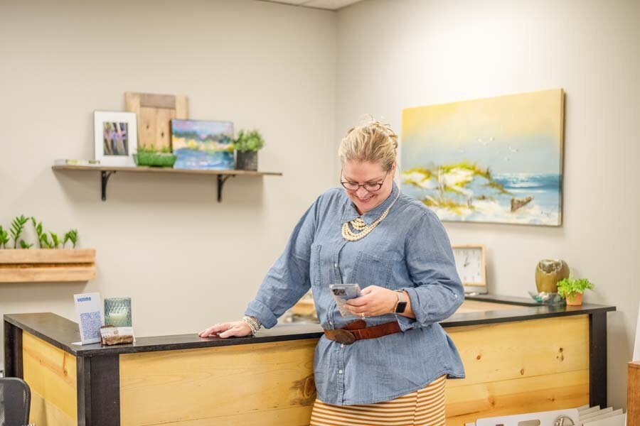 A woman in a blue shirt and striped skirt smiles while checking her smartphone at a reception desk in an office with paintings on the wall.