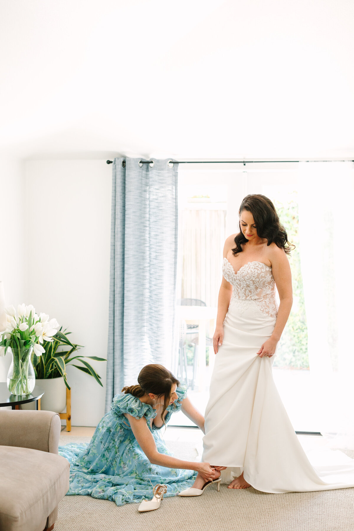 maid of honor helping bride put on shoes in a sunlit room before napa wedding ceremony.