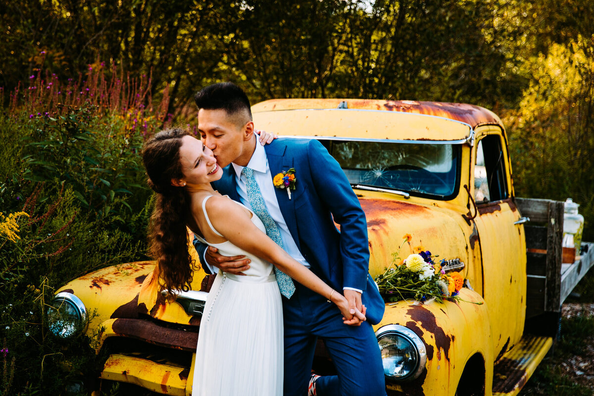 photo of a bride and groom kissing in front of an old yellow car in rural new york